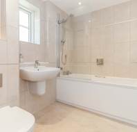 Modern bathroom with beige tiles, a wall-mounted sink, a toilet, a bathtub with a glass shower screen, and a window providing natural light.