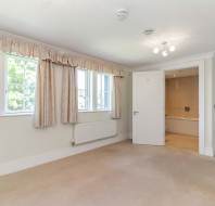 Bright, empty bedroom with beige carpet, cream walls, three windows with curtains, a radiator, and an open door leading to a bathroom.