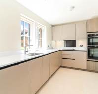 Modern kitchen with beige cabinets, integrated appliances, two ovens, a sink under two windows, and light-colored flooring.