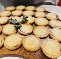 A wooden tray filled with round, golden mince pies dusted with powdered sugar, garnished with a small sprig of holly.