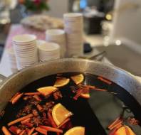 A pot of mulled wine with orange slices, cinnamon sticks, and star anise; stacks of white cups are in the background.