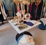 A boutique features racks of clothing, a marble table displaying accessories and jewelry, and folded scarves on blue velvet stools in a stylish, well-lit room.