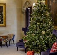 A decorated Christmas tree with wrapped gifts underneath stands in an elegant room with portraits, armchairs, and a small table with chairs.
