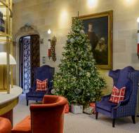 A decorated Christmas tree with wrapped gifts underneath stands between two blue armchairs in a room with beige walls, red chairs, and a portrait hanging above.