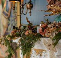 A decorated white mantle with pine branches, dried flowers, a gold bow, and a candle, beneath a wall sconce and framed artwork.
