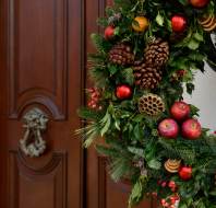 A festive wreath made of pinecones, apples, berries, and greenery hangs on a wall next to a wooden door with a decorative metal knocker.
