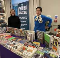 Two women stand behind a table displaying various books at an indoor bookshop event, with a "The Cobham Bookshop" banner in the background.