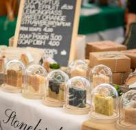 A market stall displays handmade soaps under glass domes, boxed products, bundled washcloths, and a chalkboard price list in the background.