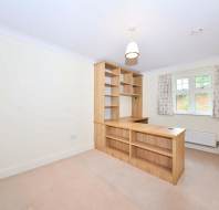 Empty room with a built-in wooden desk and shelving unit, a large window with curtains, and a ceiling light fixture. Walls are light-colored, and the floor is carpeted.