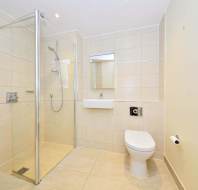 Modern bathroom with beige tiles, featuring a glass-enclosed shower, wall-mounted sink, toilet, small mirror, and a stainless steel towel heater. Grab bar and toilet paper holder installed.