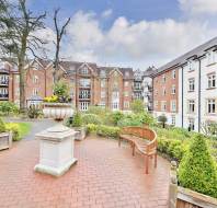 A courtyard with a brick path, benches, a large planter with flowers, and residential buildings in the background. Cloudy sky overhead.