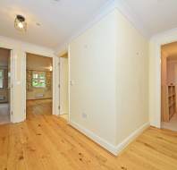 A hallway with light wooden floors leads to multiple rooms. The doors are open, revealing a bedroom with carpet, a lit bathroom, and another room. Walls are painted light yellow.