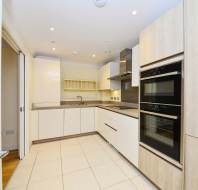 A modern kitchen with light wood cabinets, two built-in ovens, a stove, sink, and overhead shelving. The floor is tiled, and recessed lighting illuminates the space.