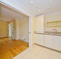 A modern kitchen with white cabinets, a sink, and a drying rack. Sliding doors open to a room with wooden floors and floral curtains.