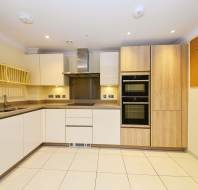 Modern kitchen with beige cabinets, stainless steel appliances, and tiled flooring. Double oven, range hood, and dish rack are visible. Bright lighting and neutral tones dominate the space.