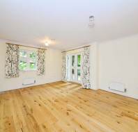 An empty room with light wood flooring, cream walls, floral curtains on windows, and double doors leading outside.