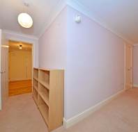 Empty room with light purple walls, beige carpet, and a wooden bookshelf. A doorway opens to a corridor with wooden flooring. Ceiling lights are present.
