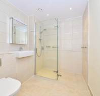 A modern bathroom with beige tiles, a wall-mounted sink, a toilet, and a glass-enclosed shower. A mirror is above the sink and a stainless steel heated towel rail is on the wall.