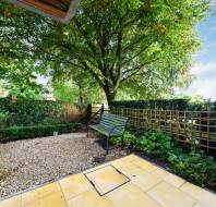 A small garden with a stone patio, bench, and tree. Fence and greenery surround the area under a clear sky.