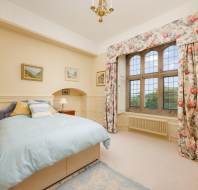 Bedroom with a large bed, floral curtains, and a bay window. Walls are cream-colored with wainscoting. Three small landscape paintings hang above the bed. Carpet is light-colored.