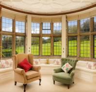 A cozy sitting area with a large bay window, armchair, and sofa. The window provides a view of a green landscape. The room features curtains and decorative pillows.