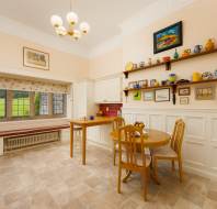 A bright kitchen with white cabinets, a wooden table and chairs, colorful decor on shelves, red tiled backsplash, and large windows overlooking a grassy area.
