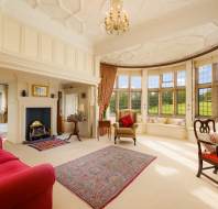 Elegant living room with large bay windows, red sofas, an ornate rug, and a fireplace. A chandelier hangs from a detailed ceiling, and the room overlooks a garden.