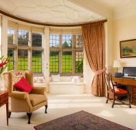 A cozy room with large bay windows, a wingback chair, a wooden desk, a vase of flowers, and a patterned rug. Sunlight streams through, illuminating the elegant decor and outside greenery.