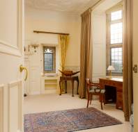 A traditional study room with a wooden desk, chair, and window with drapes. The space is carpeted and features cream-colored walls and a small lamp on the desk.
