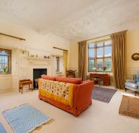 A cozy living room with a red sofa, patterned armchairs, and a fireplace. Large windows with curtains let in natural light. Various rugs cover the carpeted floor.