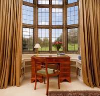 A wooden desk with a chair is set against a bay window with a view of a garden. The desk has a lamp, books, and a vase with red flowers. The room has beige curtains and a decorative rug.