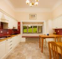 A traditional kitchen with red tile backsplash, wooden countertops, white cabinets, and a dining table. A window overlooks a grassy landscape. A chandelier and various decorations are present.