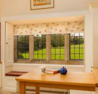 A kitchen with wooden countertops, red tiled backsplash, and a large window overlooking a green lawn. A blue teapot, a mug, and a book sit on the table.