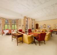 Spacious room with ornate ceiling, large windows, floral curtains, and antique furniture, including red and yellow sofas and armchairs. Walls adorned with framed pictures and a fireplace on the side.