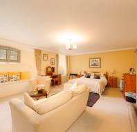 Spacious bedroom with a double bed, cream sofas, a wall-mounted TV, fireplace, and a window nook with seating. Warm yellow and beige tones dominate the room decor.