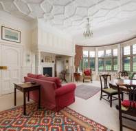 Elegant living room with ornate ceiling, red sofa, armchairs, large bay window, and decorative rugs. Classic wall moldings and a chandelier add to the sophisticated ambiance.