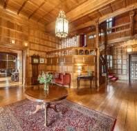 A spacious wooden-paneled room with elegant furniture, a round table with a flower vase on a rug, and ornate chandelier, leading to other rooms and a staircase.