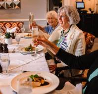 A group of elderly women sit at a dining table, each holding a glass of champagne, with plates of food and water glasses in front of them.