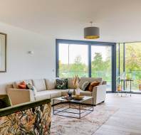 A bright living room with a beige sectional sofa, floral armchair, and glass doors leading to a balcony. Decor includes a framed artwork, patterned rug, and various cushions.