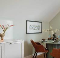 A cozy dining area with a round table set for four, two brown chairs, and minimalist decor. Nearby, a countertop holds a wooden tray with a French press and mugs. Neutral tones dominate the space.