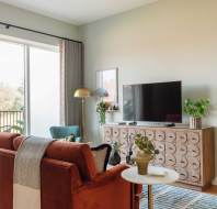 A modern living room features a red sofa, a round side table, and a TV on a cabinet adorned with plants. Large windows provide natural light, and framed artwork decorates the walls.