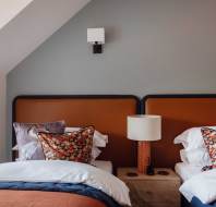 A bedroom with two single beds, each with white and colorful pillows, shares a wooden nightstand with a lamp. The walls are painted gray, and there's a wall light above the beds.