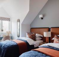 A bedroom with two twin beds featuring blue quilts and orange accents. A desk with a chair is positioned by the window, and there is a wall mirror beside it.