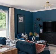 Living room with blue walls, a large window, a TV on a black stand, blue sofas with cushions, a wooden desk, and modern lamps.