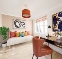 Bright room with a white sofa, colorful cushions, wooden desk, and orange chair. Decor includes abstract art, a plant, and flowers. Soft lighting from a window and orange ceiling lamp.