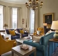 A stylish living room with armchairs, sofas, a chandelier, and large windows. Walls are adorned with paintings, and a patterned carpet covers the floor. Decorative lamps are on side tables.