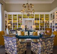 Elegant library room with a chandelier, large yellow bookshelves, a green sofa, patterned armchairs, and a central coffee table, creating a cozy reading area.