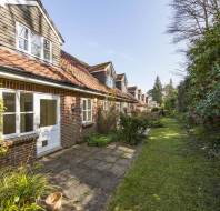 A brick house with dormer windows, white-framed doors and windows, a tiled roof, and a small patio area facing a grassy, tree-lined garden.