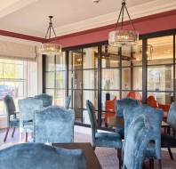 A dining room with blue upholstered chairs and glass tables. Large windows and glass doors allow natural light. Elegant chandeliers hang from the ceiling.