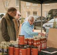 Three older adults browse jars and cans at an outdoor market stall, with buildings and a black van visible in the background.
